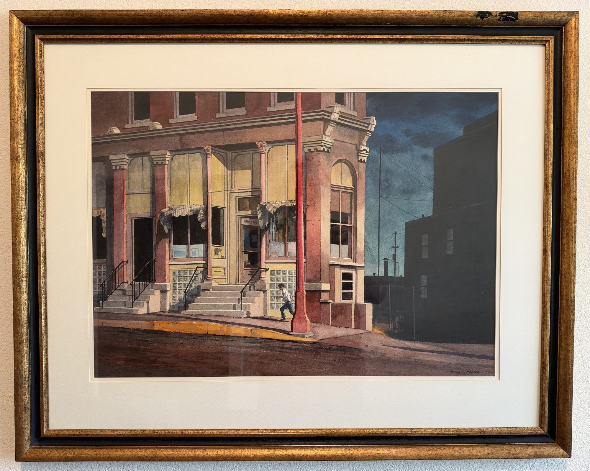 Late Evening by Floyd D. Hopper, a watercolor of a red-brick corner building with pale yellow windows and stone steps at dusk, a boy running past a red pole, dark blue sky at right, in a gilt frame with a cream mat.